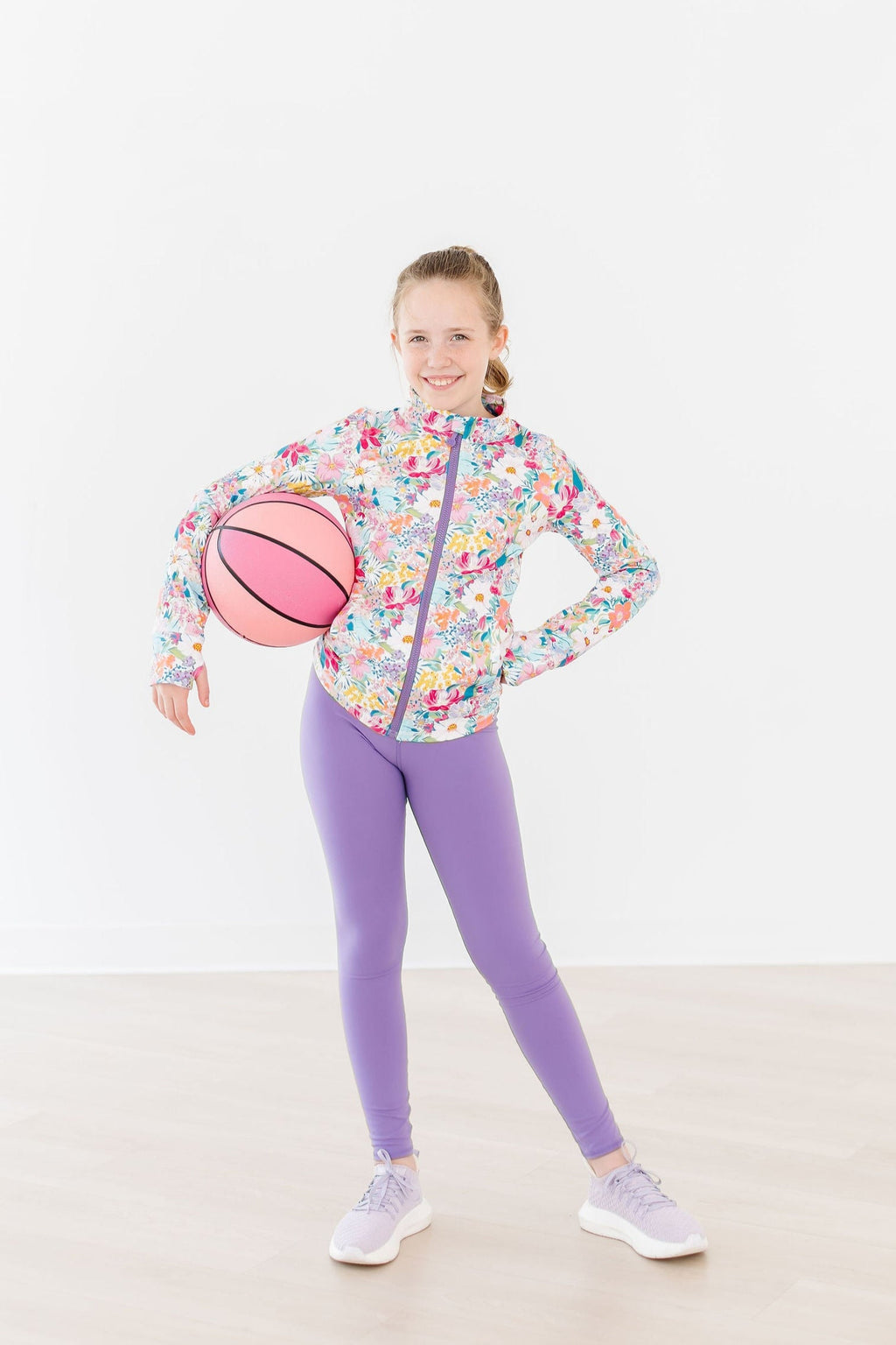 A young girl showcases the Petal Power Full-Zip Ruffle Active Jacket while posing confidently with a pink basketball, embodying a trendy and sporty look.