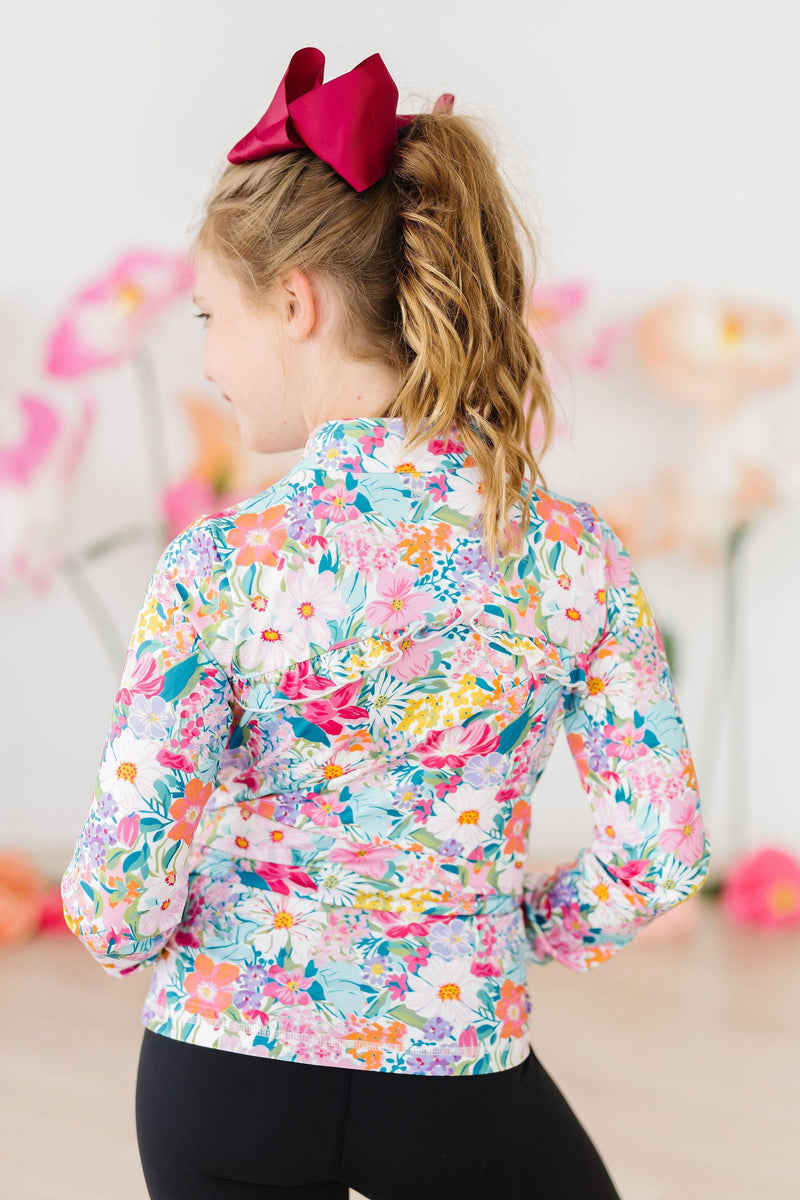 Back view of the Petal Power Full-Zip Ruffle Active Jacket, displaying vibrant floral prints and charming ruffle detailing, as the girl stands confidently.