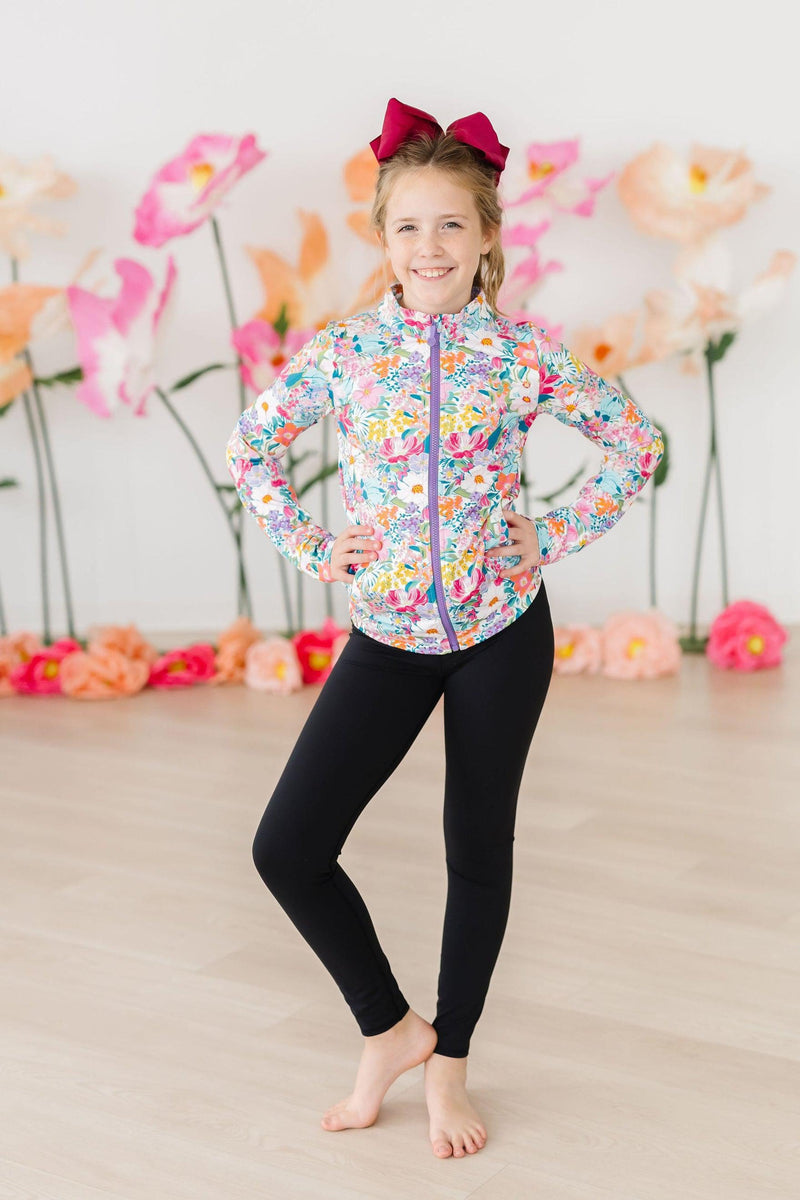 Young girl in the Petal Power Full-Zip Ruffle Active Jacket smiling brightly, paired with sleek black leggings, ready for fun and fitness!