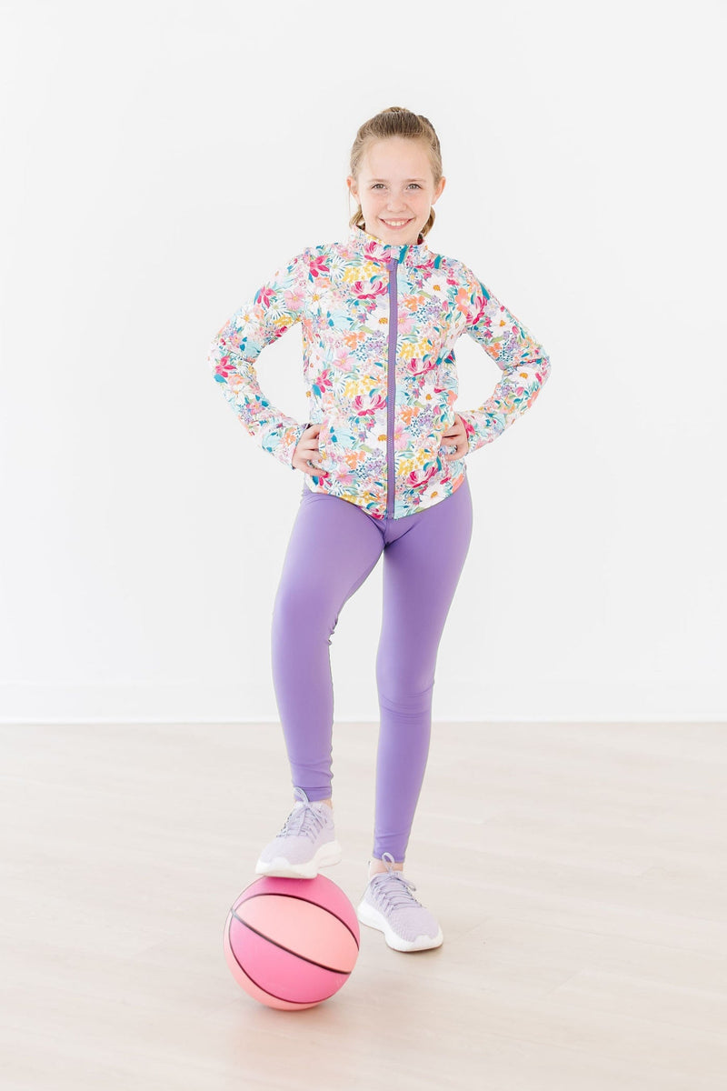 Girl striking a pose in the Petal Power Full-Zip Ruffle Active Jacket and purple leggings, holding a pink basketball, radiating style and sportiness.