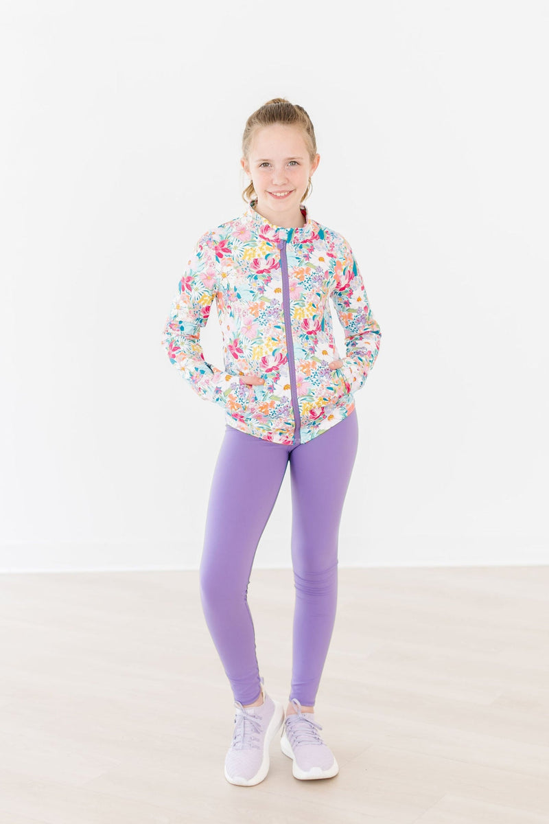 Stylish girl wearing the Petal Power Full-Zip Ruffle Active Jacket from Mila & Rose ®, posing confidently in vibrant floral patterns and purple leggings, perfect for both play and practice.