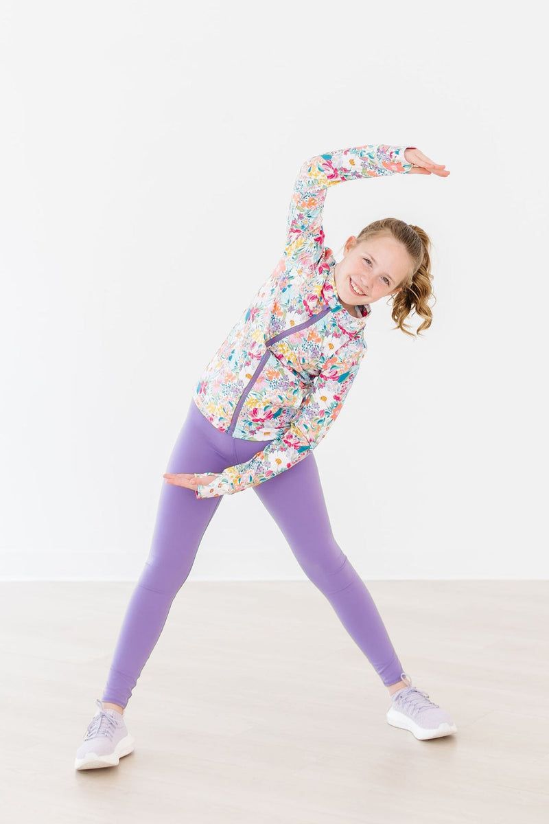 A girl performing a stretch in her Petal Power Full-Zip Ruffle Active Jacket, demonstrating flexibility and flair during her workout.