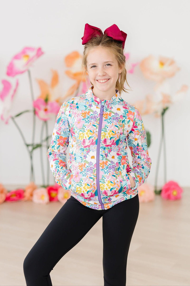 A cheerful girl poses in her Petal Power Full-Zip Ruffle Active Jacket with a bow in her hair, surrounded by colorful flowers, ready for fun!