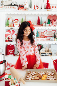 Girl in pink peppermint long sleeve tutu dress with red bow baking snowflake cookies amidst Christmas decor.
