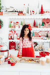 Girl in pink peppermint long sleeve tutu dress with red skirt and bow, baking Christmas cookies in a festive kitchen.
