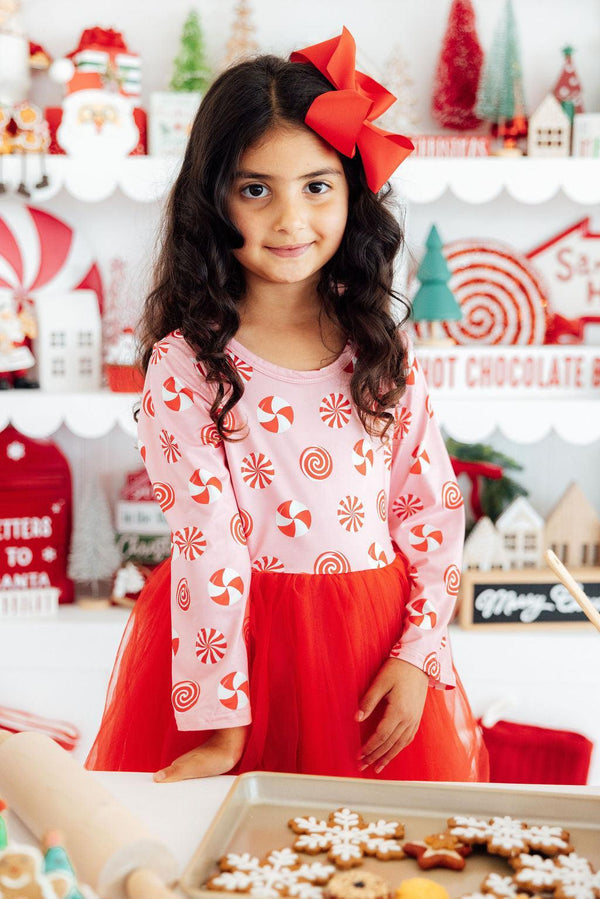 Girl in pink peppermint long sleeve tutu dress with red bow, standing by holiday cookies.