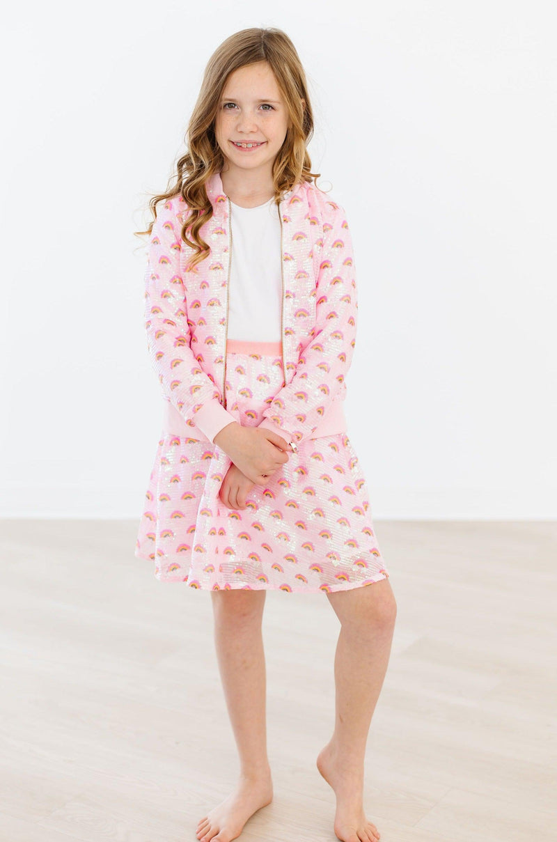 A girl in a Pink Rainbow Sequin Jacket by Mila & Rose ®, standing gracefully with her arms crossed, exuding elegance and style.