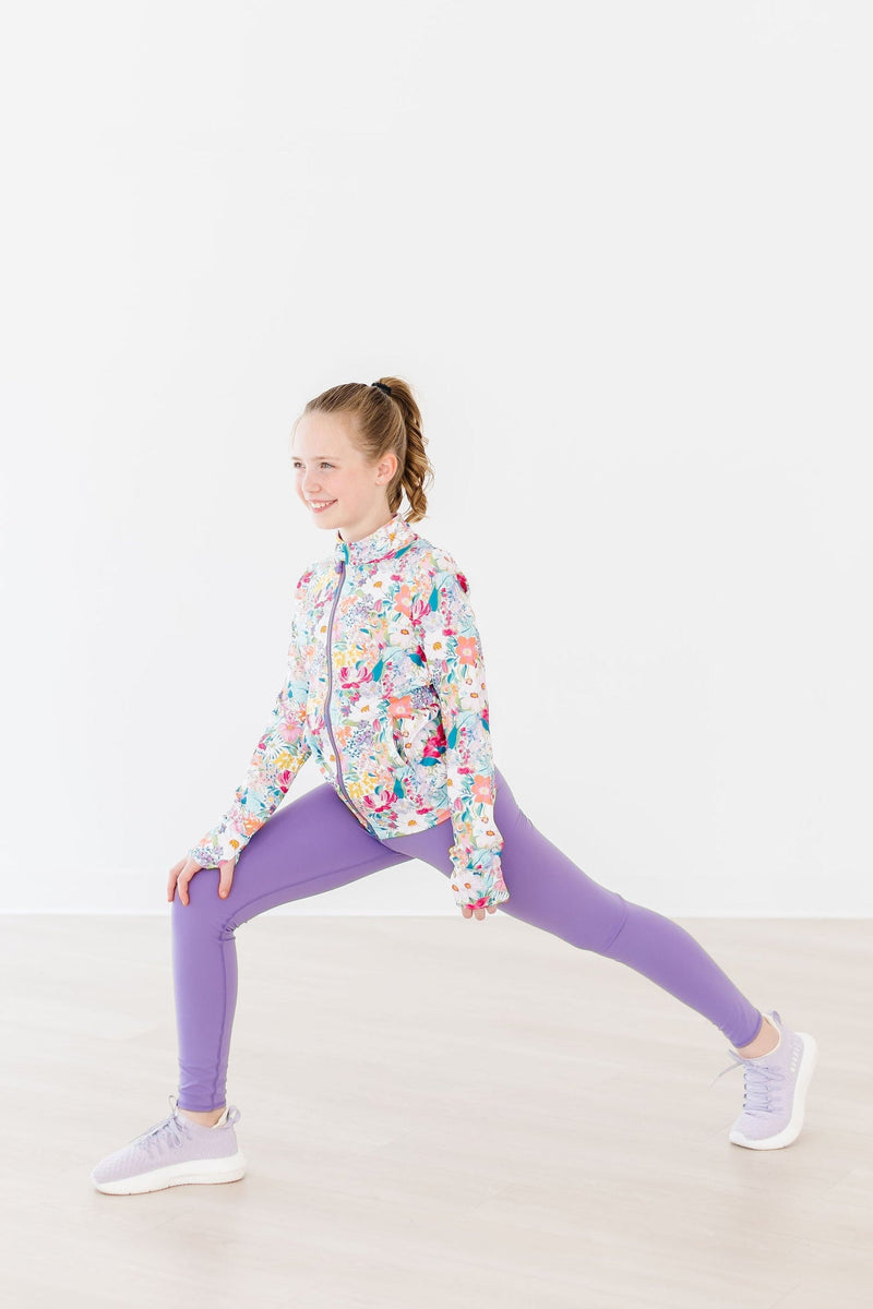 A girl wearing stylish purple leggings for girls, performing a lunge exercise in a colorful floral zip-up jacket.