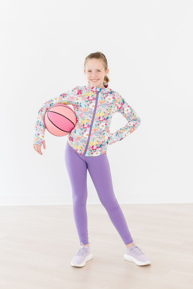 A young girl stands confidently in playful purple leggings for girls, holding a basketball, dressed in a bright floral jacket.