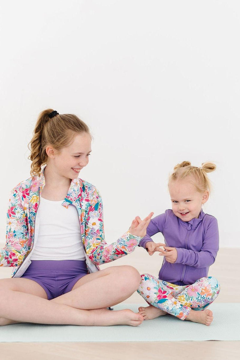 Playful scene of two girls, one in a Playful Purple Full-Zip Ruffle Girl's Active Jacket, sharing a joyful moment while dressed in vibrant activewear.