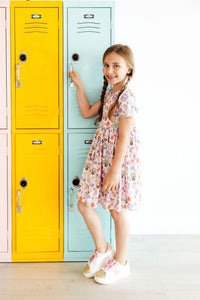 Girl wearing Playground Princess short-sleeve twirl dress with pockets, vehicle print, beside colorful lockers.