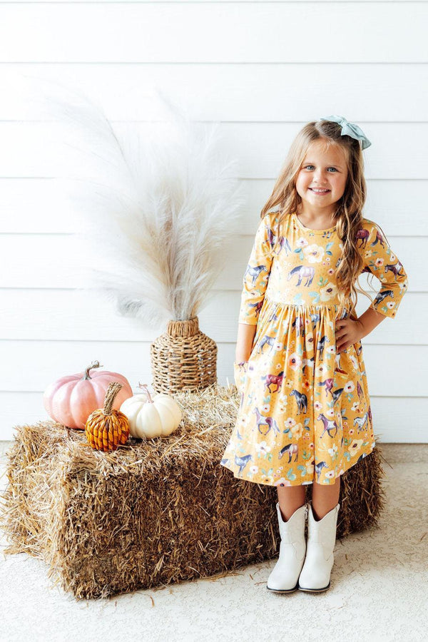 A young girl stands on a hay bale next to pumpkins and a vase of pampas grass, wearing a yellow long-sleeve dress with a colorful fall leaf pattern and white cowboy boots.