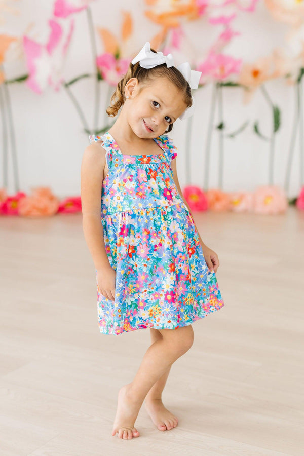 Little girl wearing a Preppy Floral Ruffle Cross Back Dress with a white bow and bright floral print.