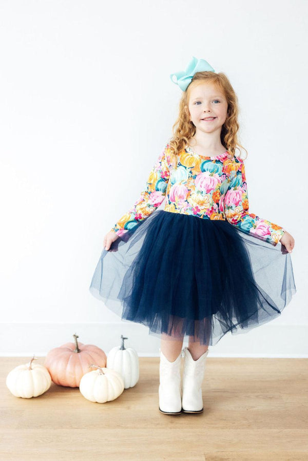Girl in colorful pumpkin print long sleeve tutu dress with navy tulle skirt and white boots, posing near pumpkins.