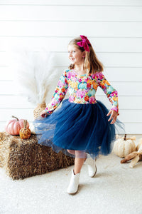 Girl twirling in a long-sleeve pumpkin print preppy patch tutu dress with navy tulle skirt, white boots, and pink bow near hay bales.