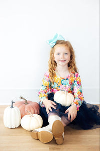 Girl in pumpkin print Preppy Patch L/S Tutu Dress with navy tulle skirt and light blue bow, sitting with decorative pumpkins.