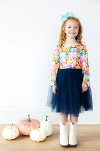 Girl wearing a preppy patch long sleeve tutu dress with colorful pumpkin print bodice and navy tulle skirt, styled with white boots and pumpkins.