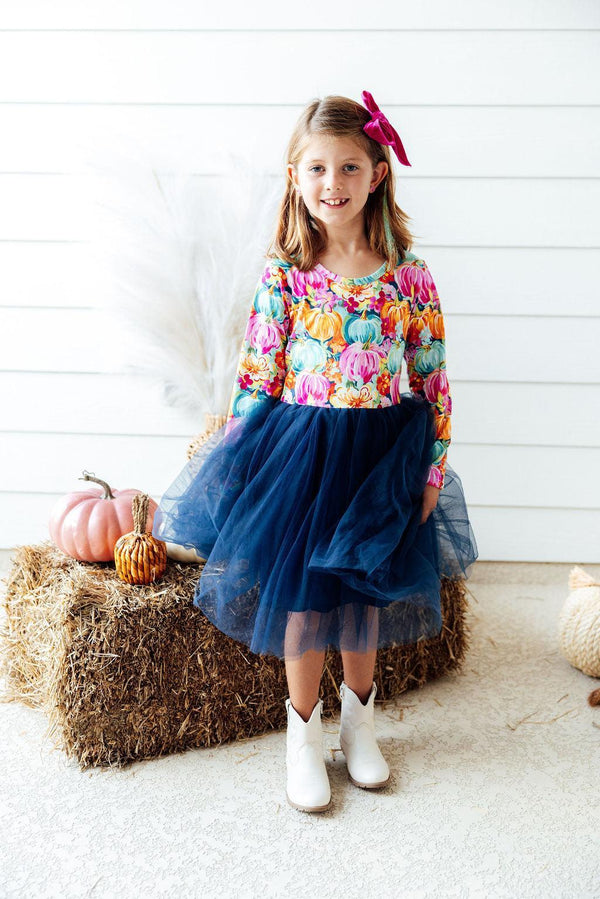 Girl wearing colorful pumpkin print long sleeve tutu dress with navy skirt and white boots near hay bale.