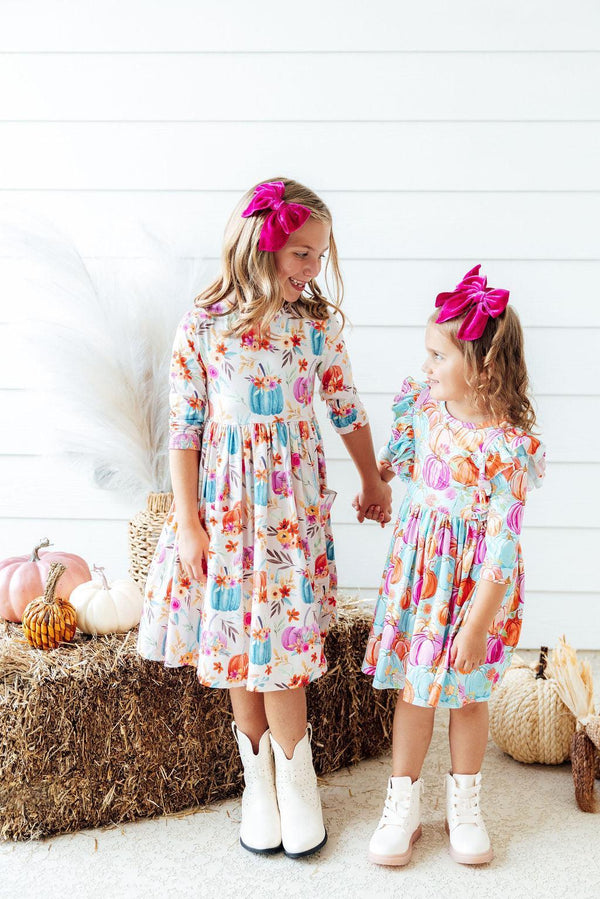 Two girls wearing Preppy Pumpkins 3/4 Sleeve Ruffle Twirl Dresses with pink bows, holding hands.