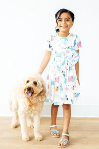 Smiling girl in a dog-print dress wears a matching puppy pals belt bag crossbody while standing next to a fluffy golden dog.