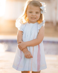 Little girl smiling in a light blue swing dress with flutter sleeves and colorful rainbow pleats down the front.