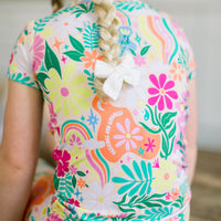 Rainbow Daisy Babe short-sleeve two-piece pajama in bright floral print, worn with braided hair and white bow.