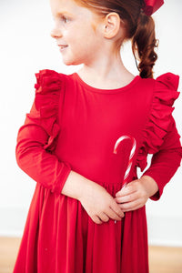 Child in red long sleeve ruffle twirl dress holding a candy cane, featuring festive ruffled shoulders for special occasions.