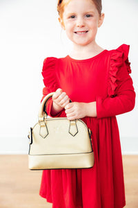 Smiling girl wearing a red long sleeve ruffle twirl dress and holding a gold handbag.