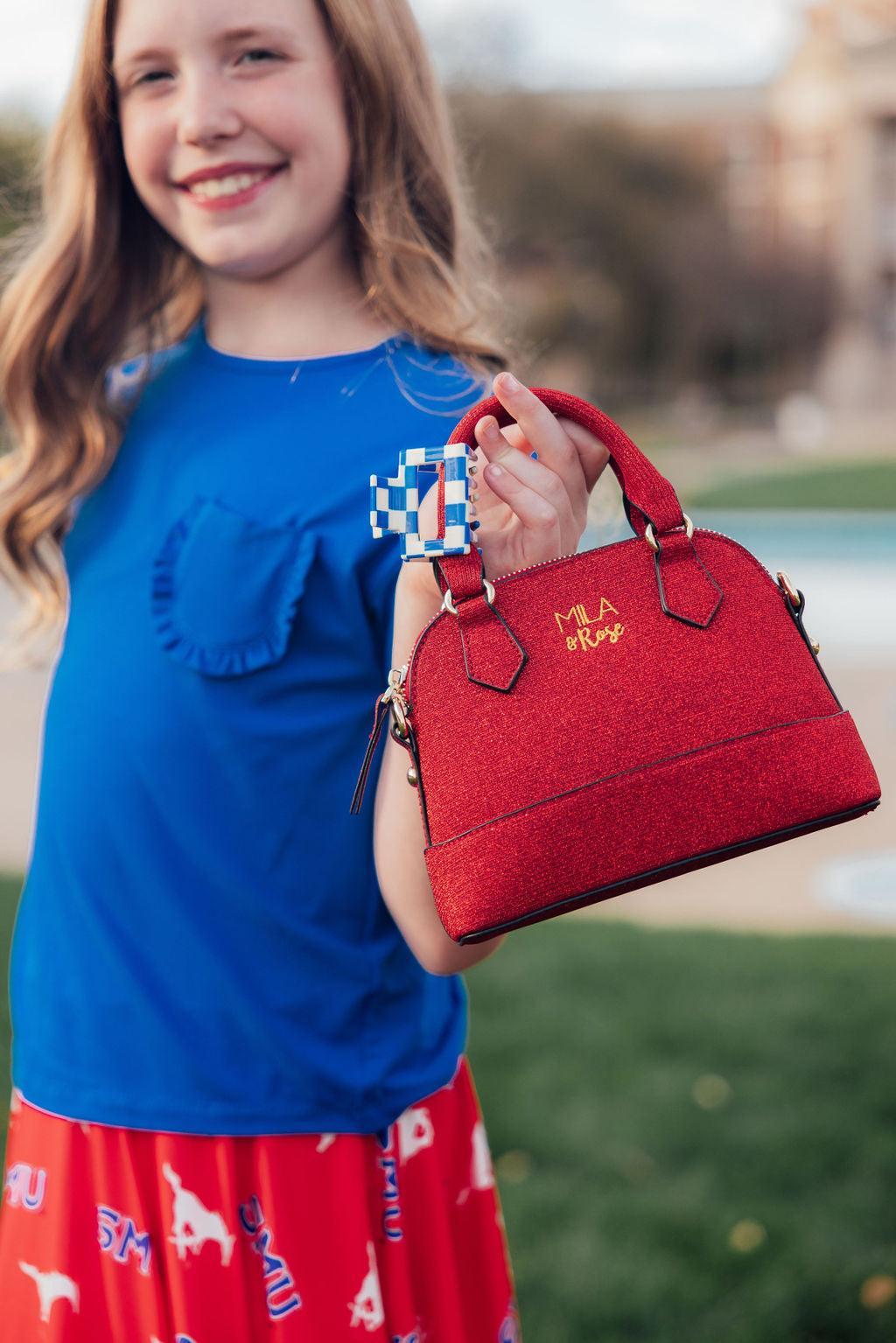 Girl holding sparkly red crossbody purse with blue checkerboard charm. Shimmer girl accessory perfect for birthdays or Christmas gifts.