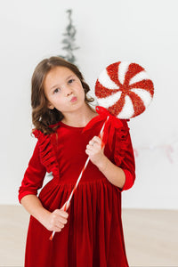 Young girl in a red velvet ruffle twirl dress holds a large glittery peppermint candy for the holidays.