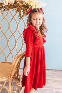 Little girl wearing a red velvet ruffle twirl dress with long sleeves and matching hair bows, standing beside a rattan chair.
