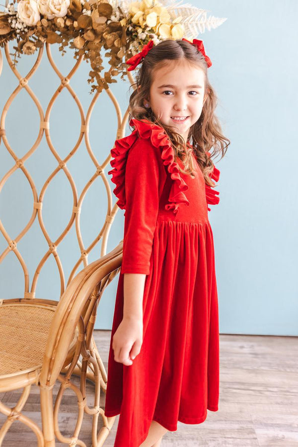 Little girl wearing a red velvet ruffle twirl dress with long sleeves and matching hair bows, standing beside a rattan chair.
