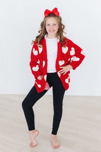 Young girl wearing a red and white coconut heart cozy cardigan with black leggings and a large red hair bow.