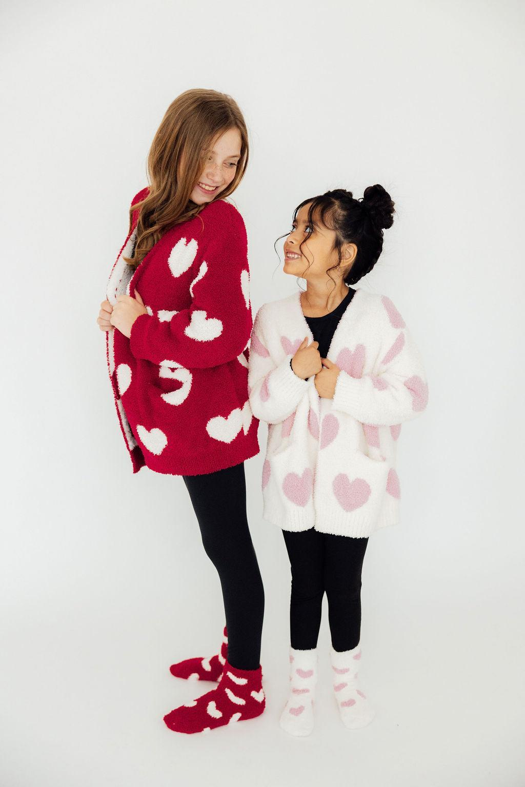 Two girls wear cozy red and white heart cardigans with matching socks, styled over black leggings for a warm, casual look.