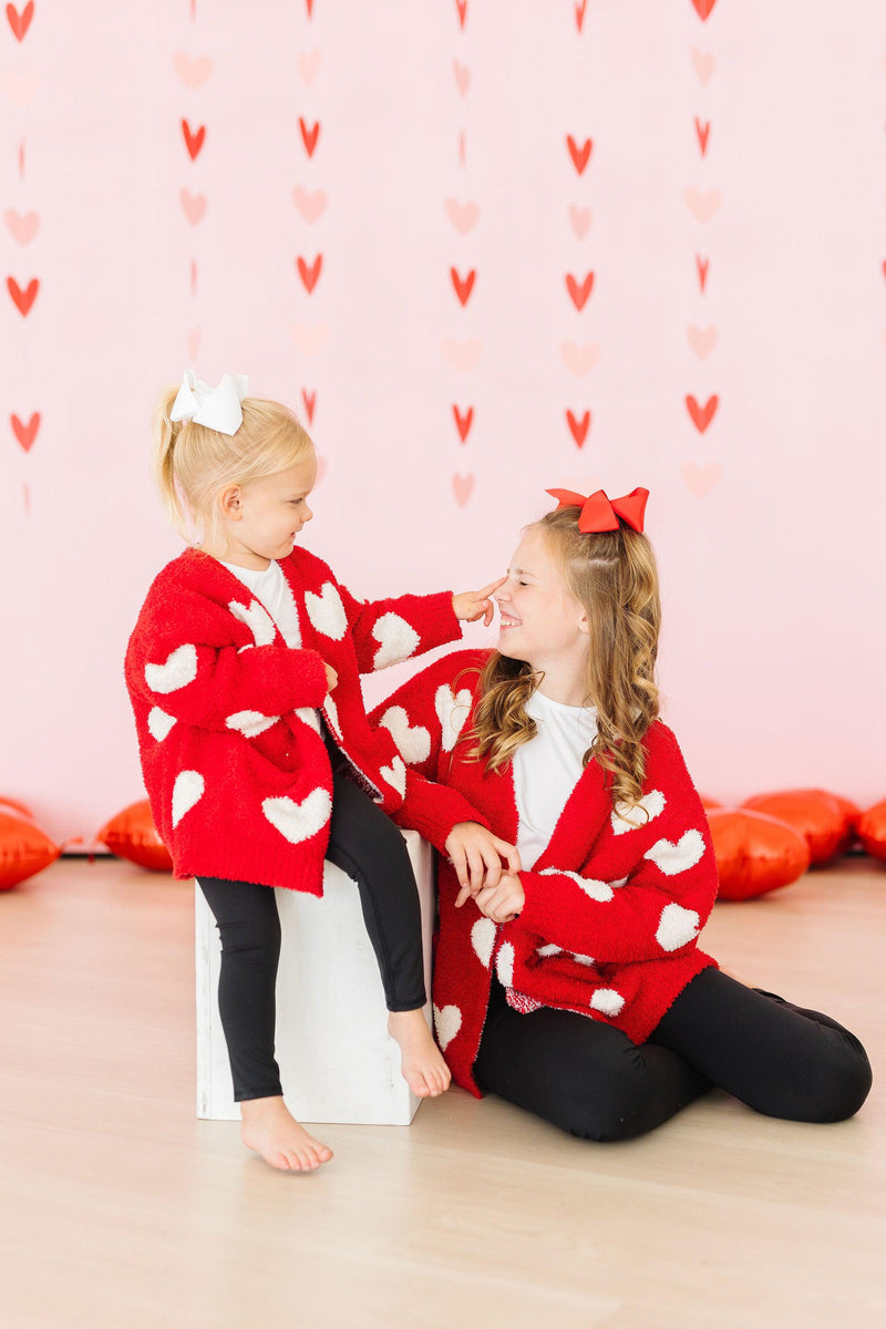 Mom and daughter wearing matching red cozy cardigans with white hearts against a pink heart backdrop.
