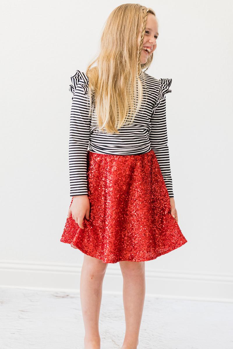 A joyful girl laughing while wearing the Red Sequin Twirl Skort from Mila & Rose, paired with a striped top. Ideal for making every moment shine, especially during celebrations.