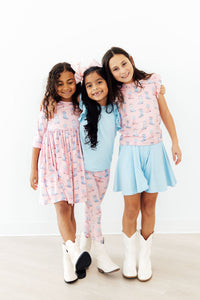 Three kids in pastel Ribbons & Rodeos outfits: pink 3/4 sleeve pocket twirl dress paired with a blue top.
