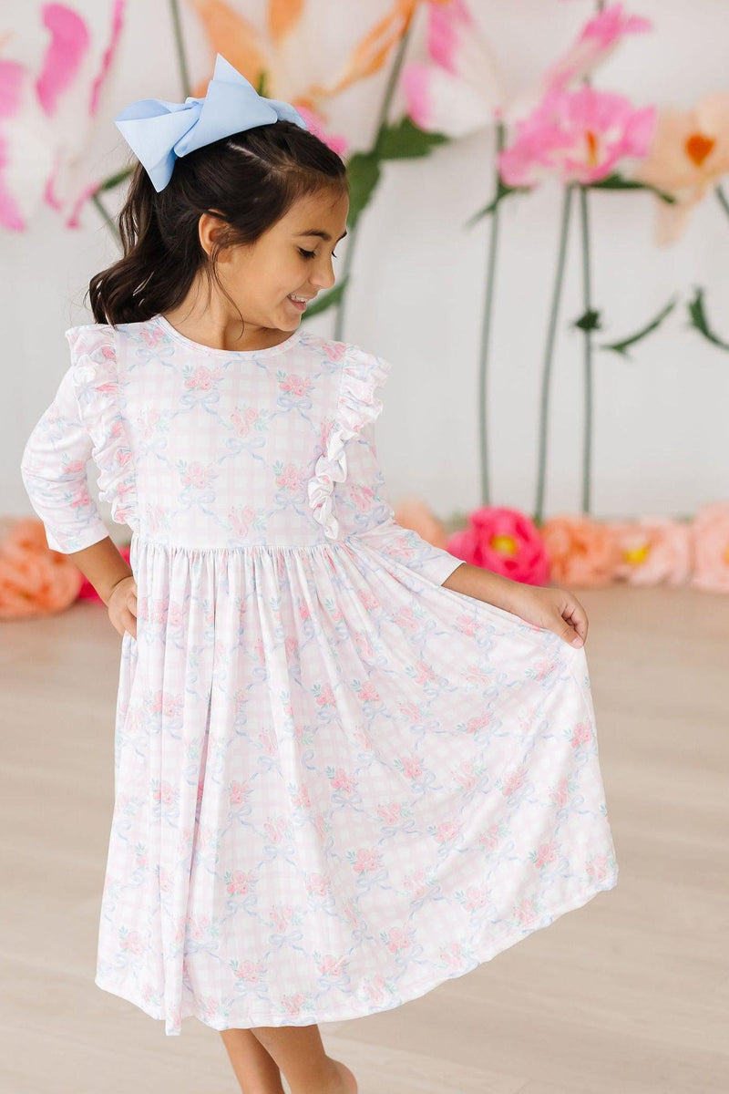 Smiling child dressed in the Ribbons & Roses 3/4 Sleeve Ruffle Twirl Dress, showcasing the dress's playful ruffle details with a big blue bow in her hair.