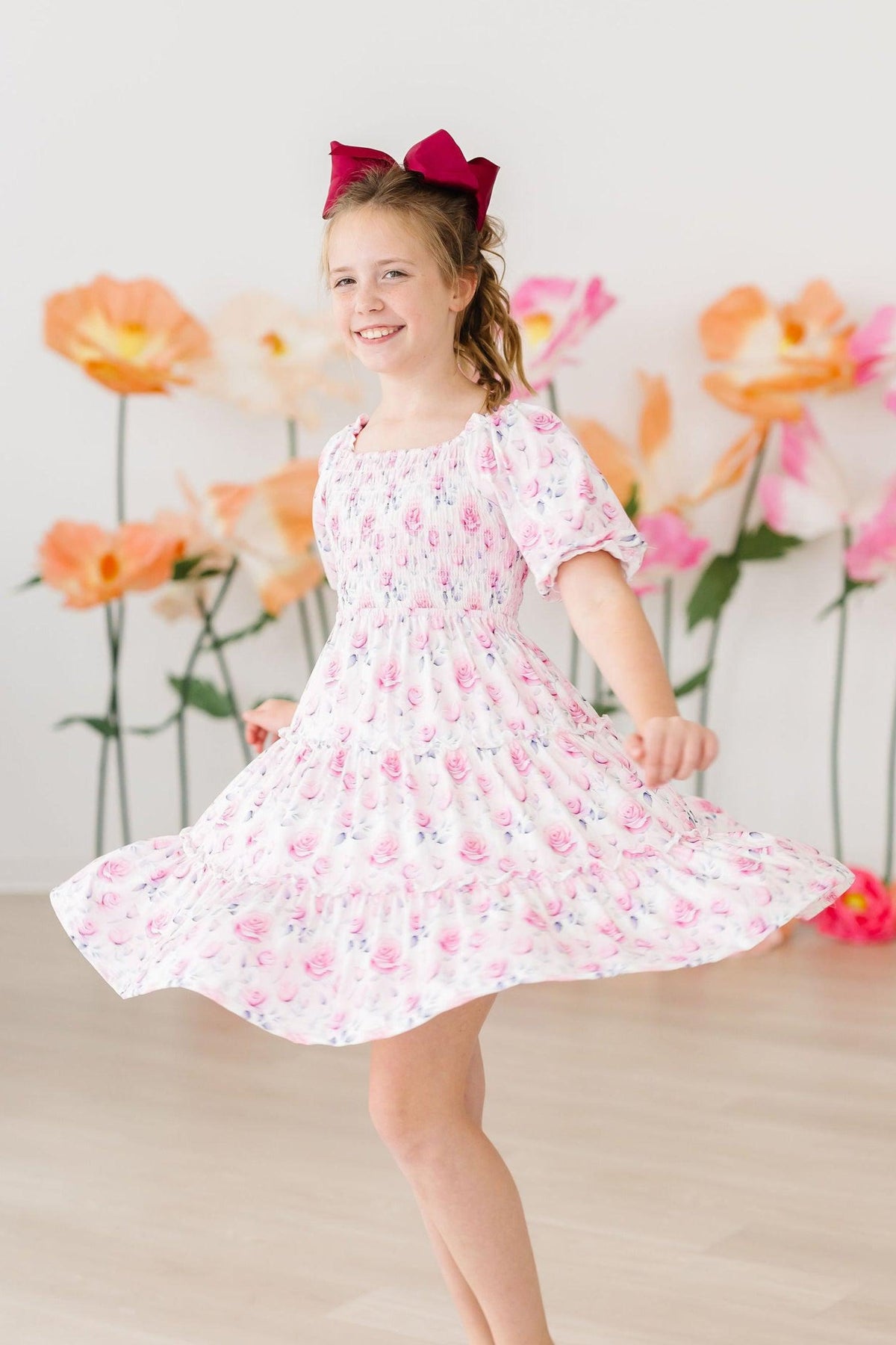 Girl twirls in Rose Garden Smocked Ruffle Dress with pink bow, floral rose print.