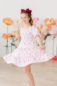 Girl twirls in Rose Garden Smocked Ruffle Dress with pink bow, floral rose print.