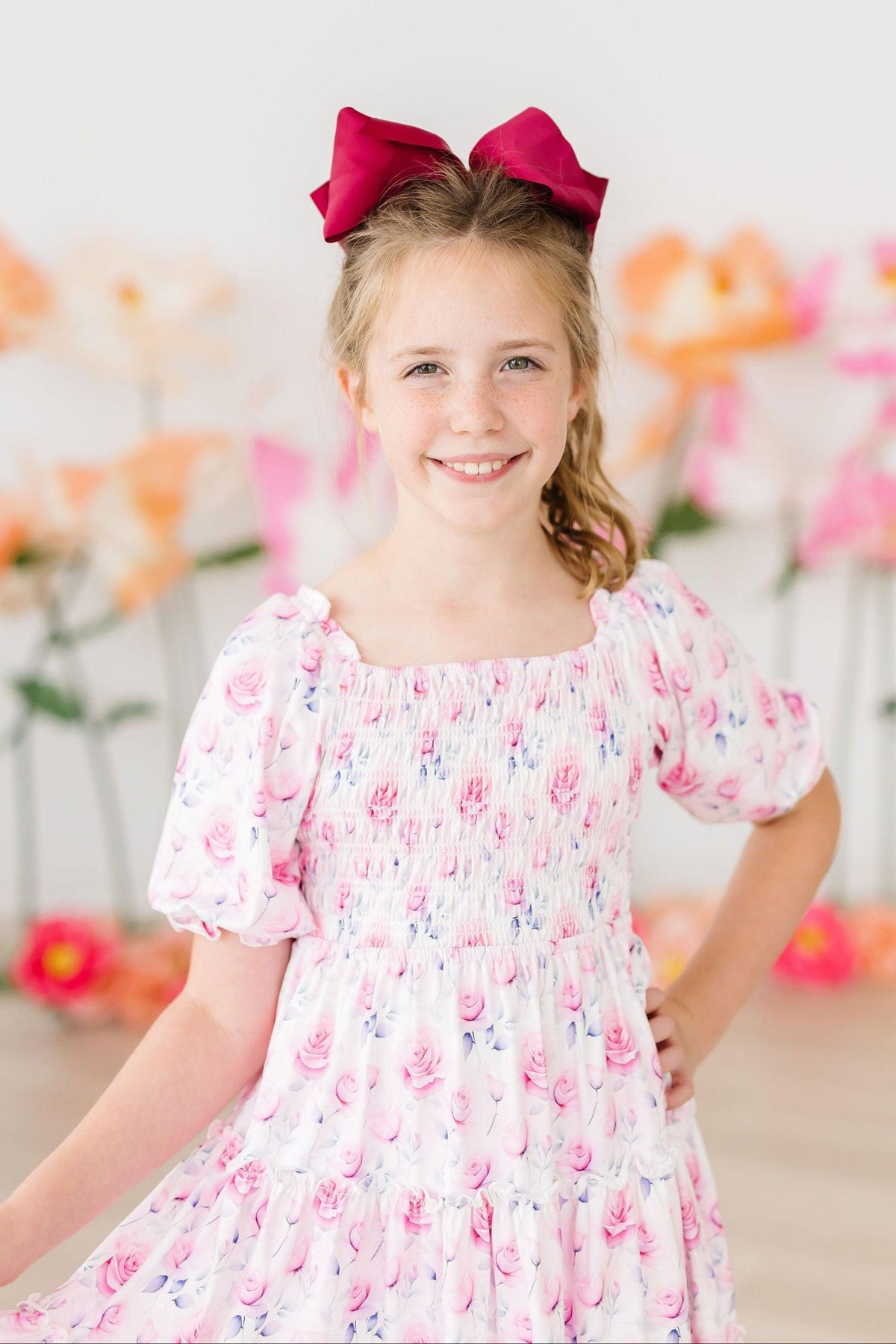 Young girl wearing Rose Garden Smocked Ruffle Dress with a pink bow, pink floral print.
