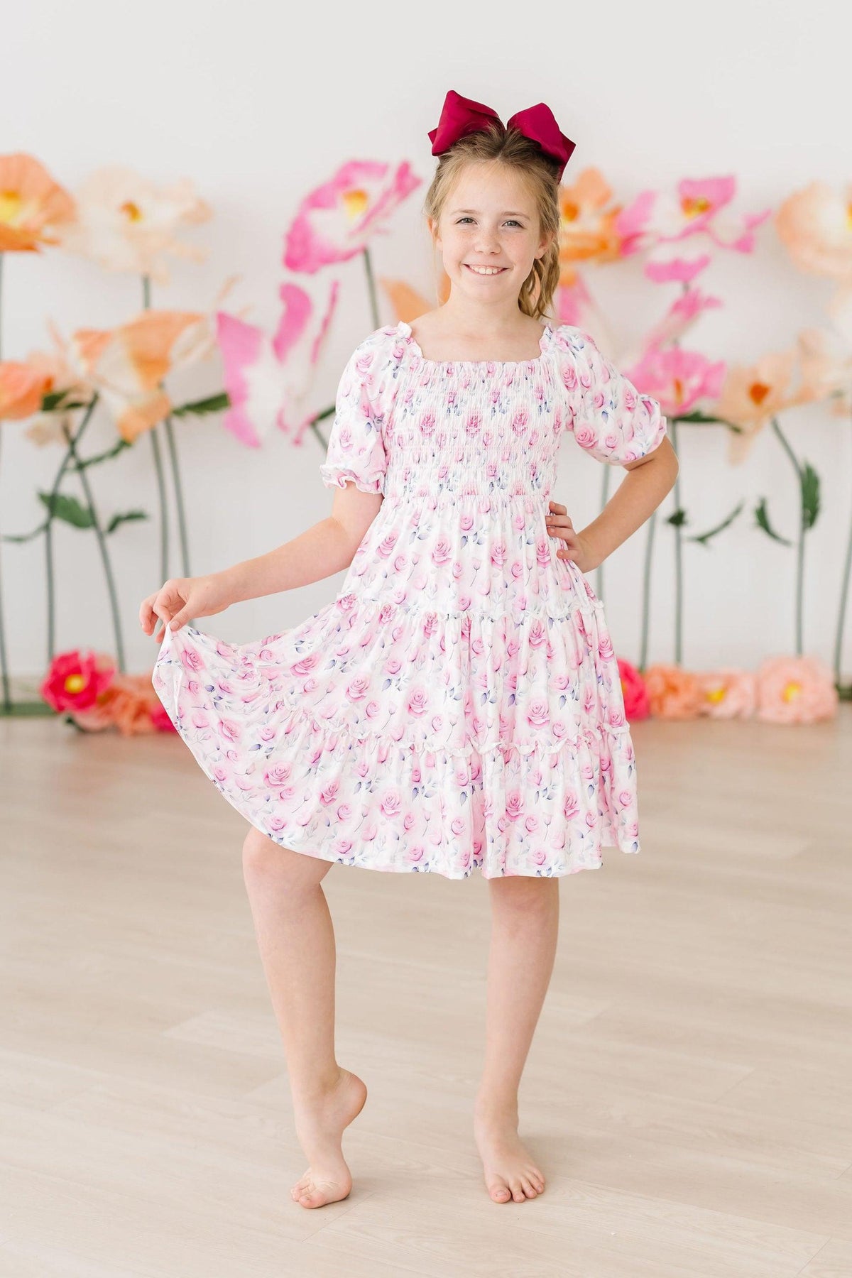Rose Garden Smocked Ruffle Dress with a smocked bodice on a smiling girl in pink floral print and maroon bow.