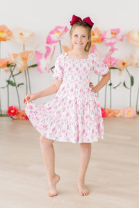 Rose Garden Smocked Ruffle Dress with a smocked bodice on a smiling girl in pink floral print and maroon bow.