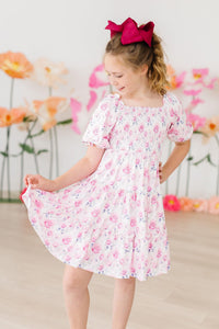 Rose Garden Smocked Ruffle Dress worn by a girl with a pink bow in a pastel floral setting.