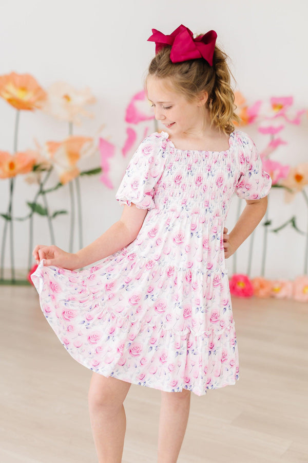 Rose Garden Smocked Ruffle Dress worn by a girl with a pink bow in a pastel floral setting.