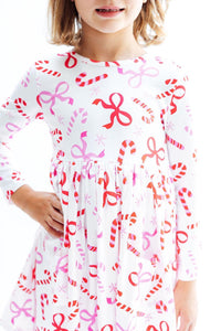 Child wearing white long sleeve twirl dress with red candy canes, pink bows, and snowflakes. Features a full circle skirt and front pocket.