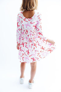 Back view of a girl twirling in a white long sleeve dress printed with red candy canes and pink bows, featuring a scoop neckline and full circle skirt.