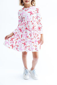 Girl twirling in a white long sleeve dress with red candy cane and pink bow print, featuring a full circle skirt and light blue sneakers.