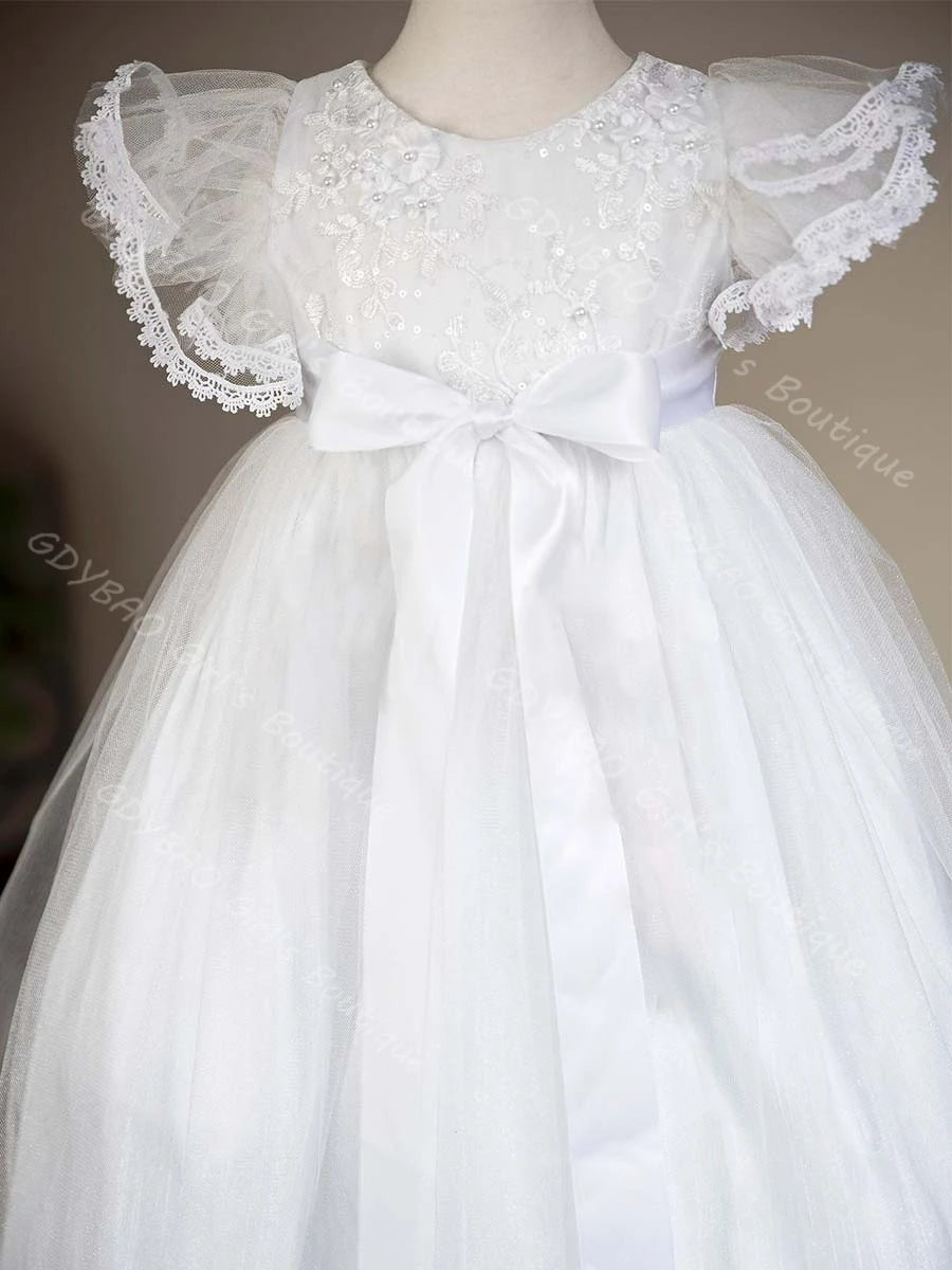 White baby girl christening gown with lace flutter sleeves, beaded floral embroidery, and satin bow sash on tulle skirt.