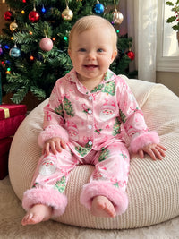 Baby girl in pink Santa print pajama set with fuzzy cuffs, sitting by a Christmas tree.